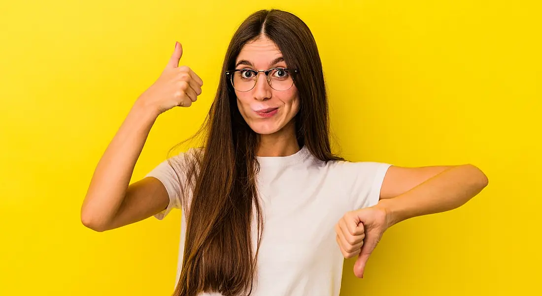 Woman doing thumbs up with one hand and thumbs down with the other as though evaluating a remote job. She is standing against a yellow background.