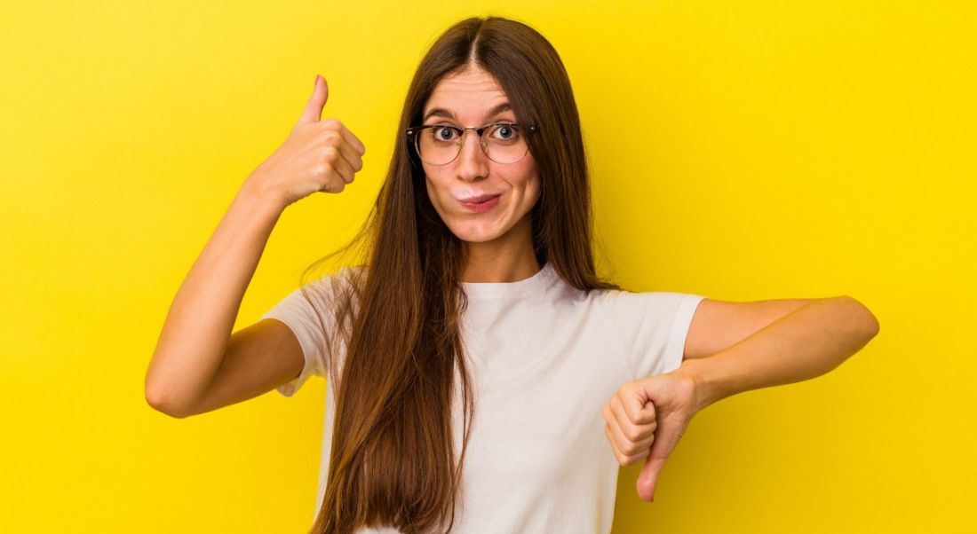 Woman doing thumbs up with one hand and thumbs down with the other as though evaluating a remote job. She is standing against a yellow background.