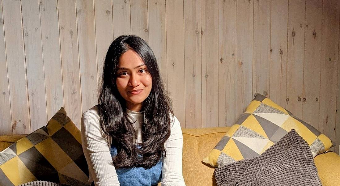 A young woman with long dark hair smiles at the camera while sitting on a yellow couch with cushions either side against a wooden wall.