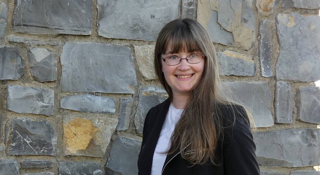 A young woman graduate with long hair smiling at the camera. She is wearing a black blazer and standing in front of a stone wall.