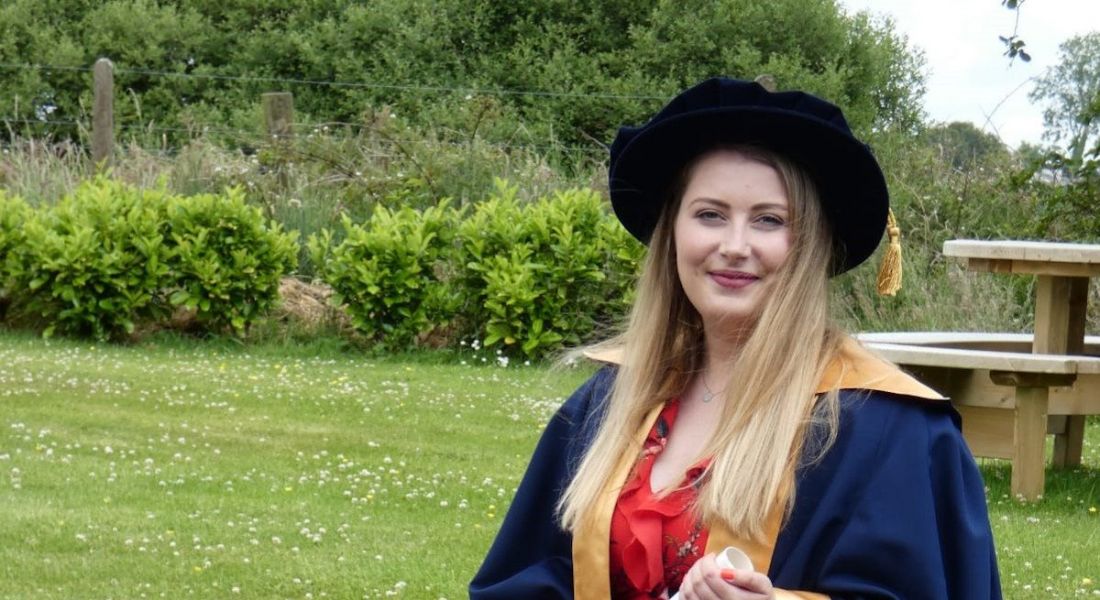A woman in a doctoral graduation cap and gown standing outside with grass behind her. She is Fiona Hennessy from MSD.