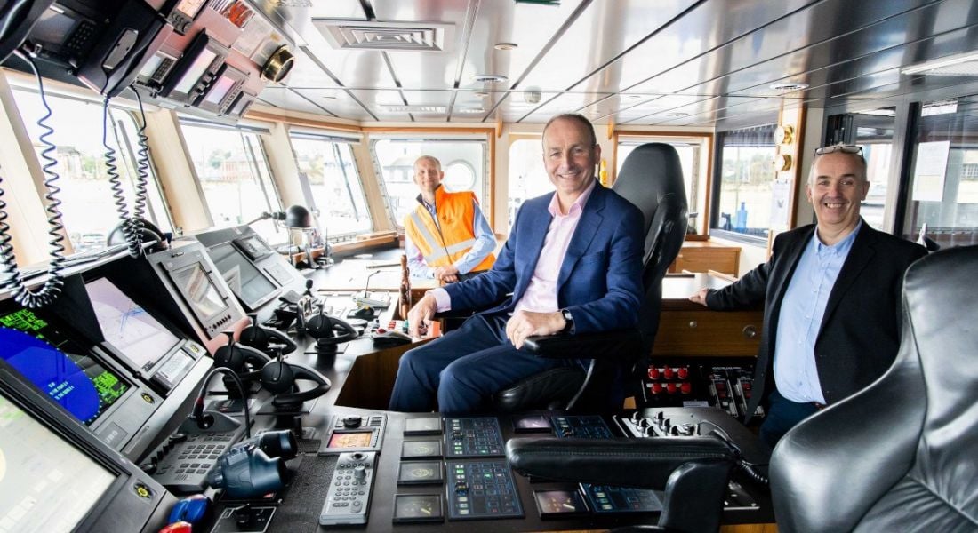 Three men sit at the helm of a boat. One man is wearing a high-vis vest while the others are in navy suits.