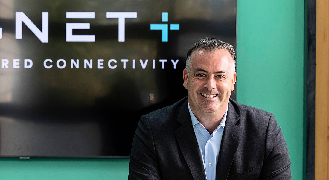 A man in a suit smiling at the camera. Behind him is a wall that shows the second half of the Magnet Plus logo.