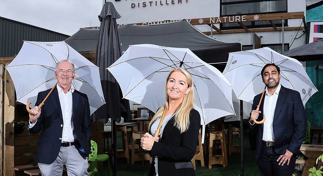 Two men and a woman in business attire stand outside a small distillery, each carrying large white umbrellas to take shelter from the rain.