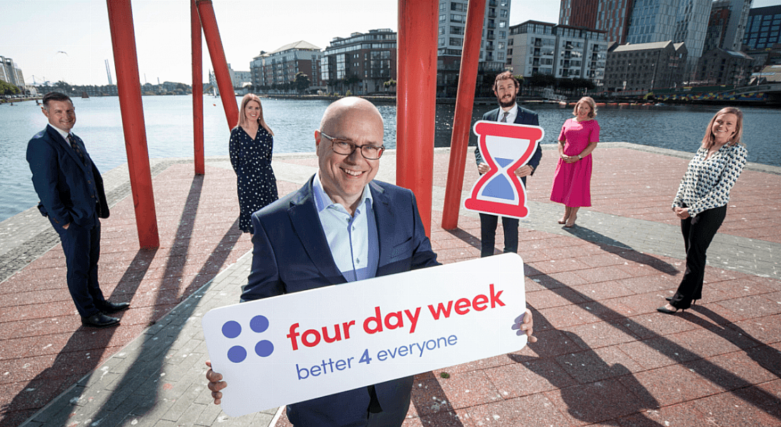 Members of the Four Day Week Ireland campaign standing outside in a formation watching one of their number holding a sign with their logo on it.