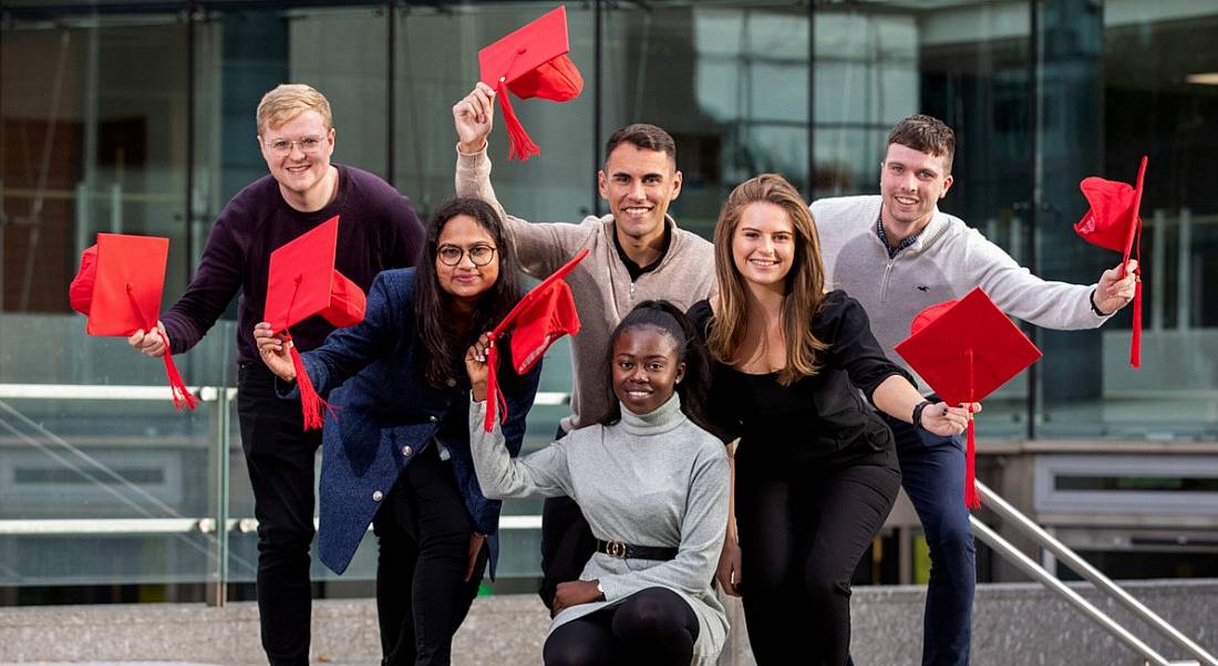 Vodafone Ireland graduate programme participants standing and kneeling in a group outside a glass building waving red graduate caps in the air.