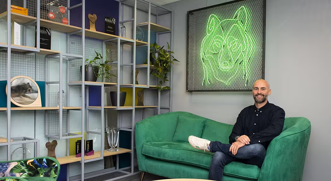 Alan Coleman of Wolfgang Digital sitting in an office reception area on a green couch under a neon green wolf sign.