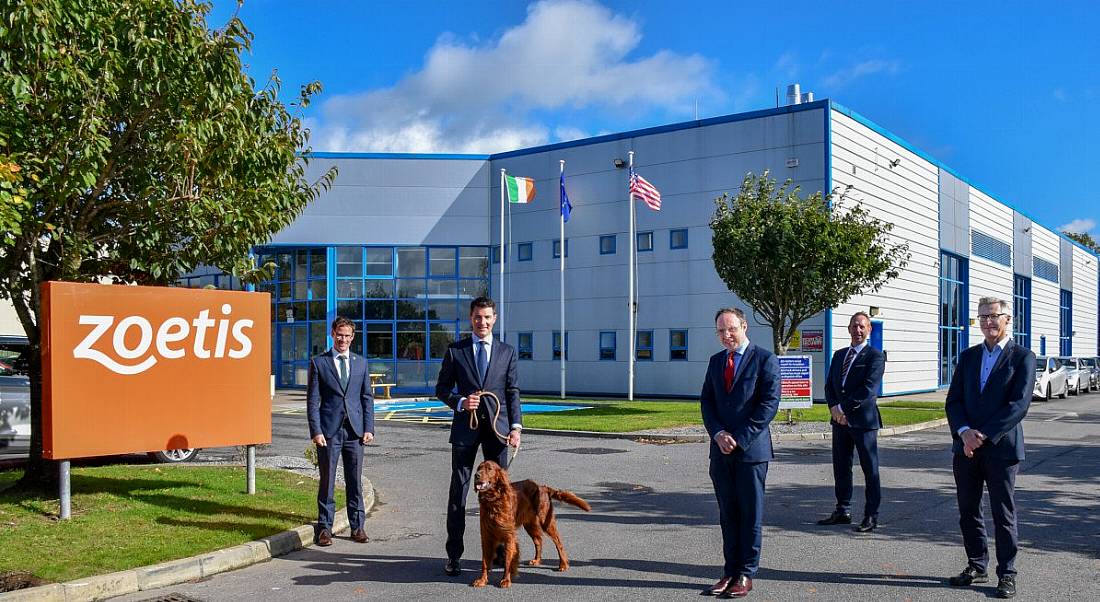 Five men in suits stand outside a large manufacturing facility along with an Irish red setter, held on a leash by one of the men.