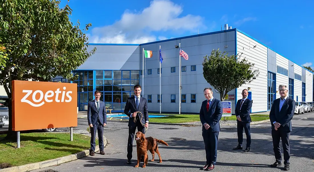 Five men in suits stand outside a large manufacturing facility along with an Irish red setter, held on a leash by one of the men.