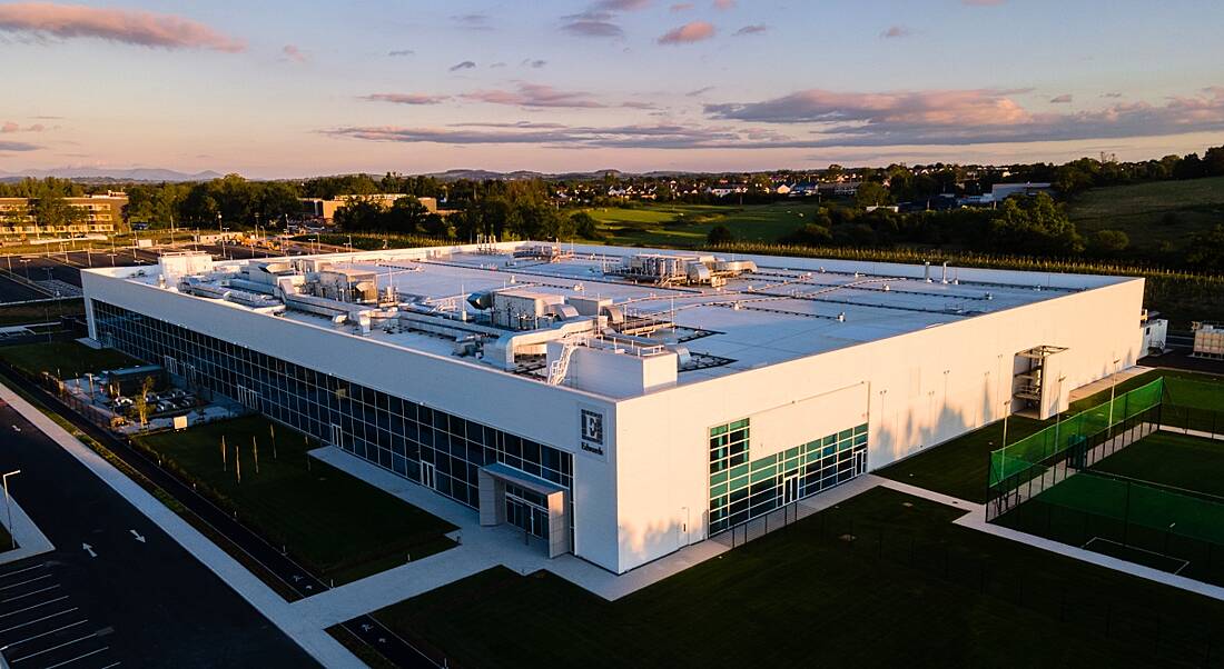 The new Edwards Lifesciences manufacturing facility in Castletroy, Limerick. It is a white, sprawling building with the company logo in front and green grass all around. Hills and trees in the background.