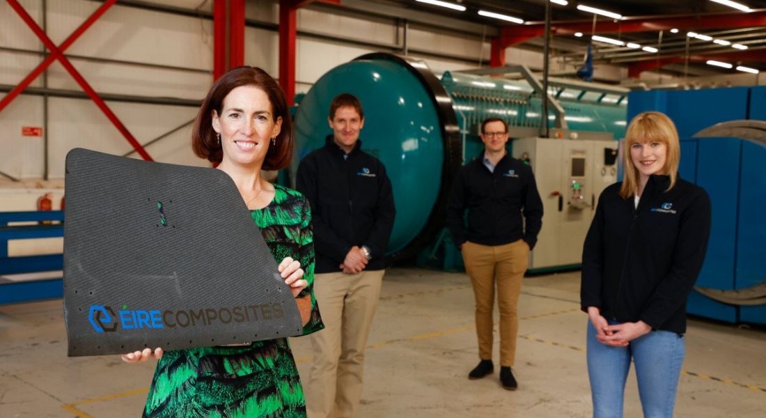 A woman in a floral dress in the foreground holds up a square structure bearing the ÉireComposites logo. Three people stand socially distanced in the background in matching company fleeces at a large manufacturing facility.
