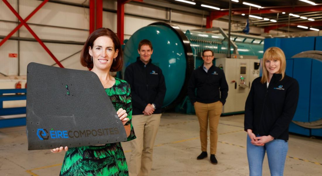 A woman in a floral dress in the foreground holds up a square structure bearing the ÉireComposites logo. Three people stand socially distanced in the background in matching company fleeces at a large manufacturing facility.