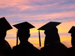 A small graduation cap sits on top of a blue pencil inside a jar beside another pencil against an out-of-focus background of greenery. It symbolises doctoral graduates.