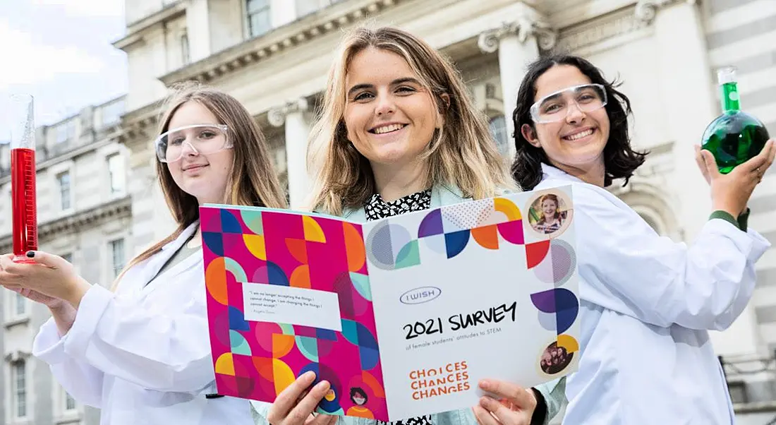 Three young women standing in a row dressed in lab gear and smiling and holding lab equipment outside at the launch of I Wish's report.