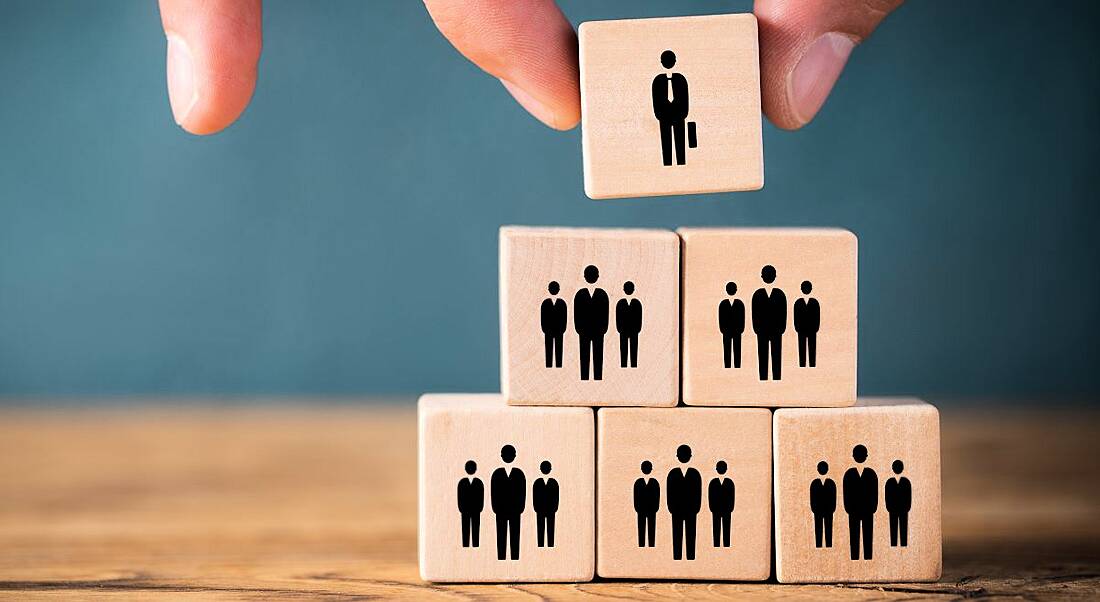 Six wooden blocks stacked in a pyramid shape with businesspeople drawn on them. The top block is being removed by a hand.