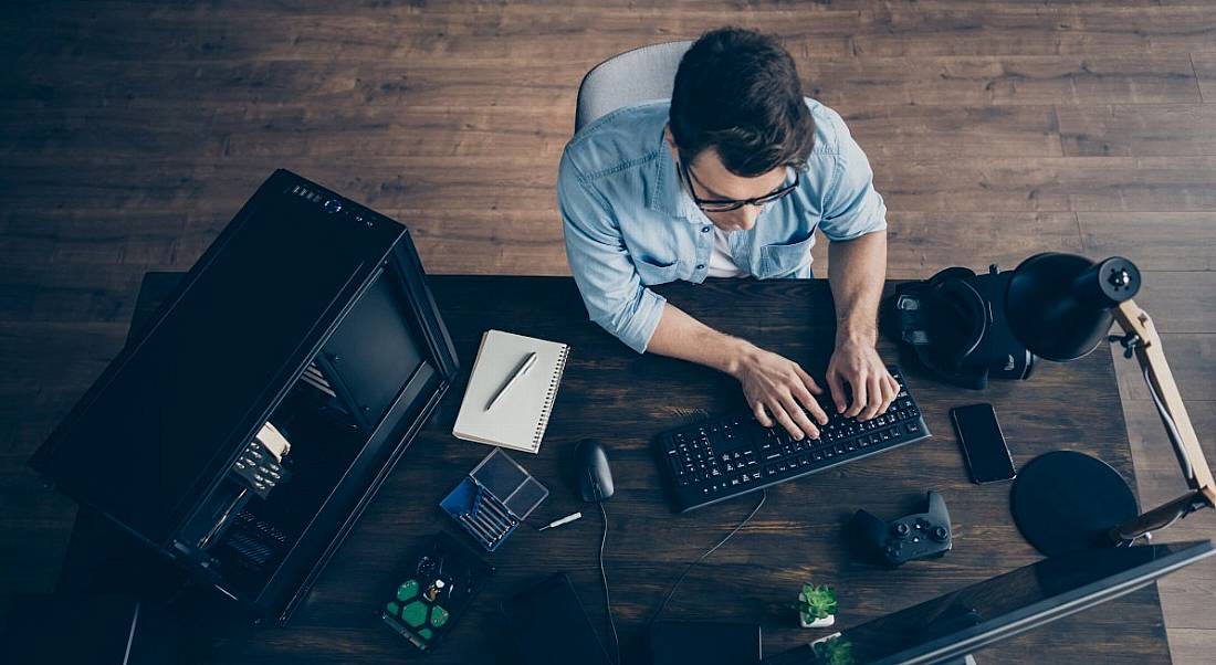 A high-angle shot looking down at a young man with glasses working at a large desk with multiple technology items around him.