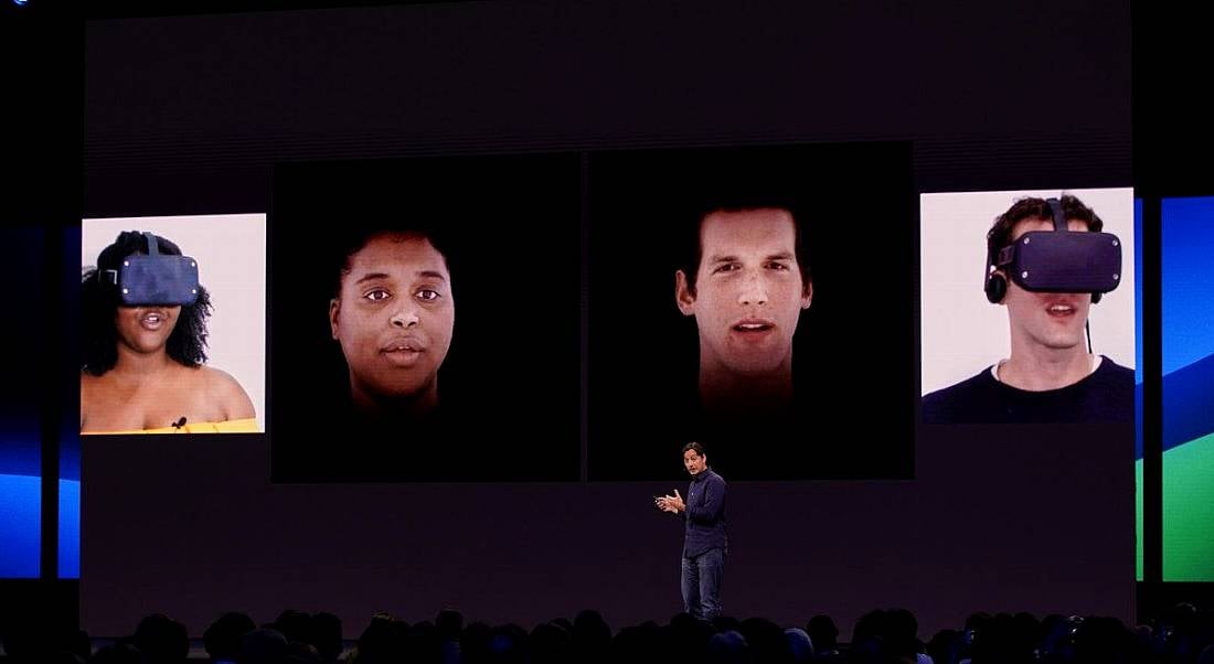 A man stands on stage in front of a large conference audience, with a supersize screen behind him demonstrating Oculus VR technology and headsets.