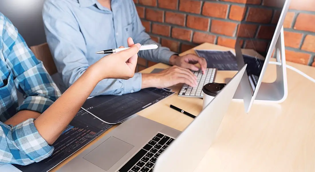 Two people sitting at a desk, working in front of a laptop and a desktop monitor.