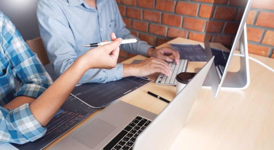 Two people sitting at a desk, working in front of a laptop and a desktop monitor.