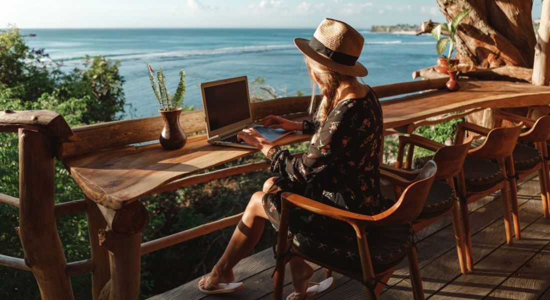 Woman remote working at a laptop sitting at a beachfront bar in the sunshine.