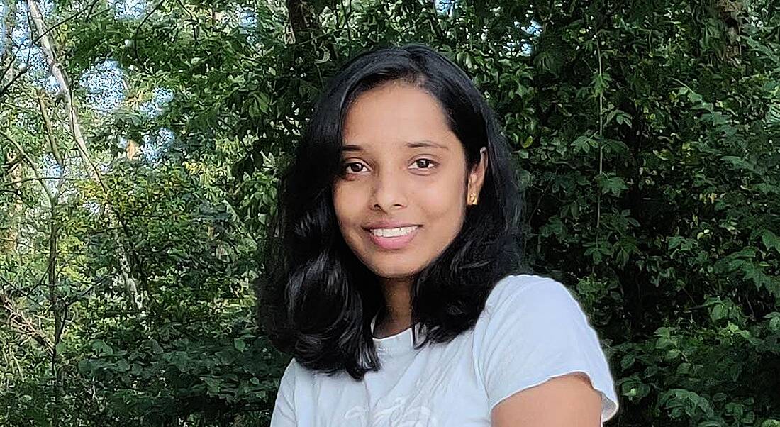 A young woman with dark hair smiles at the camera against a backdrop of trees and greenery.