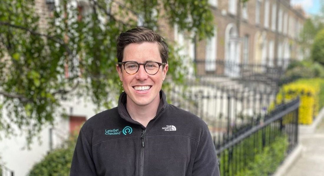 A man with glasses smiles at the camera while standing on a residential street with a row of houses behind him. His fleece has the LetsGetChecked logo on it.