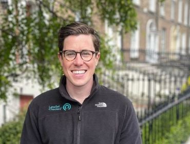 A man with glasses smiles at the camera while standing on a residential street with a row of houses behind him. His fleece has the LetsGetChecked logo on it.