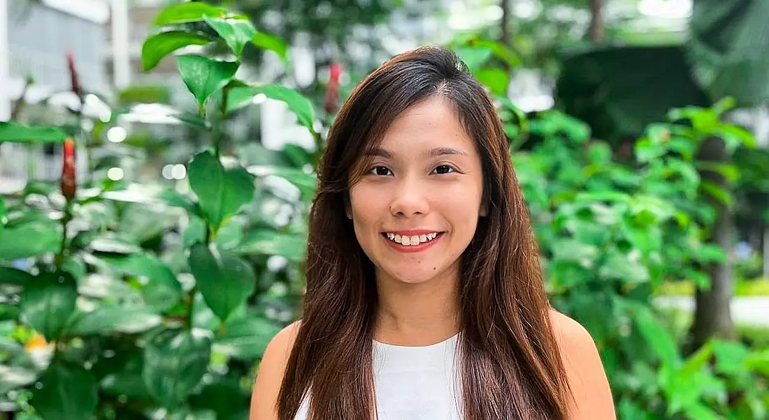 A young woman with long brown hair smiling at the camera against a backdrop of leafy plants.