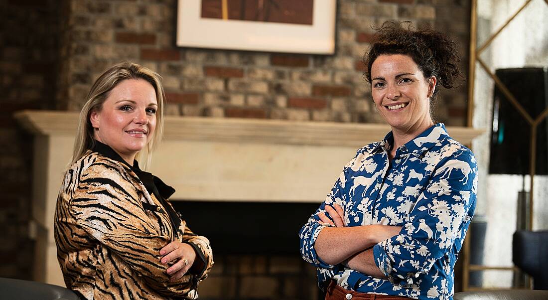 Two women stand with their arms folded in a cosy room.