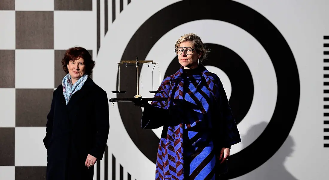 Two women stand against a black and white patterned wall. The woman on the right is holding a gold scales.