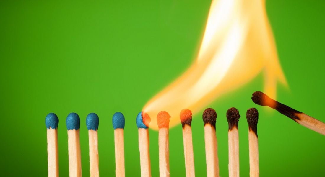 A line of matches against a green background. One has lit the start of the line and a large flame is burning through them one by one, symbolising burnout.