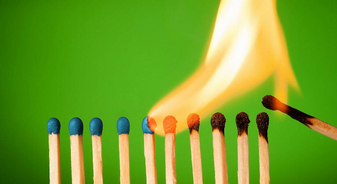 A line of matches against a green background. One has lit the start of the line and a large flame is burning through them one by one, symbolising burnout.