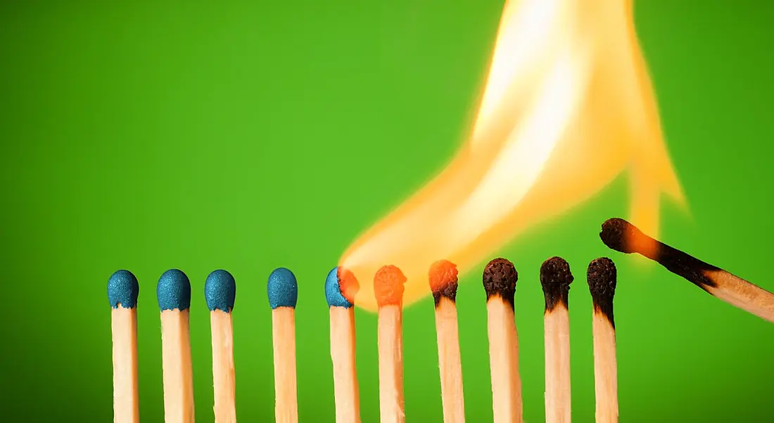 A line of matches against a green background. One has lit the start of the line and a large flame is burning through them one by one, symbolising burnout.