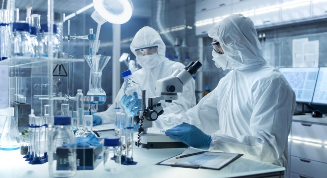 Two scientists in full protective gear work in a lab with a microscope and other biopharma paraphernalia in front of them.
