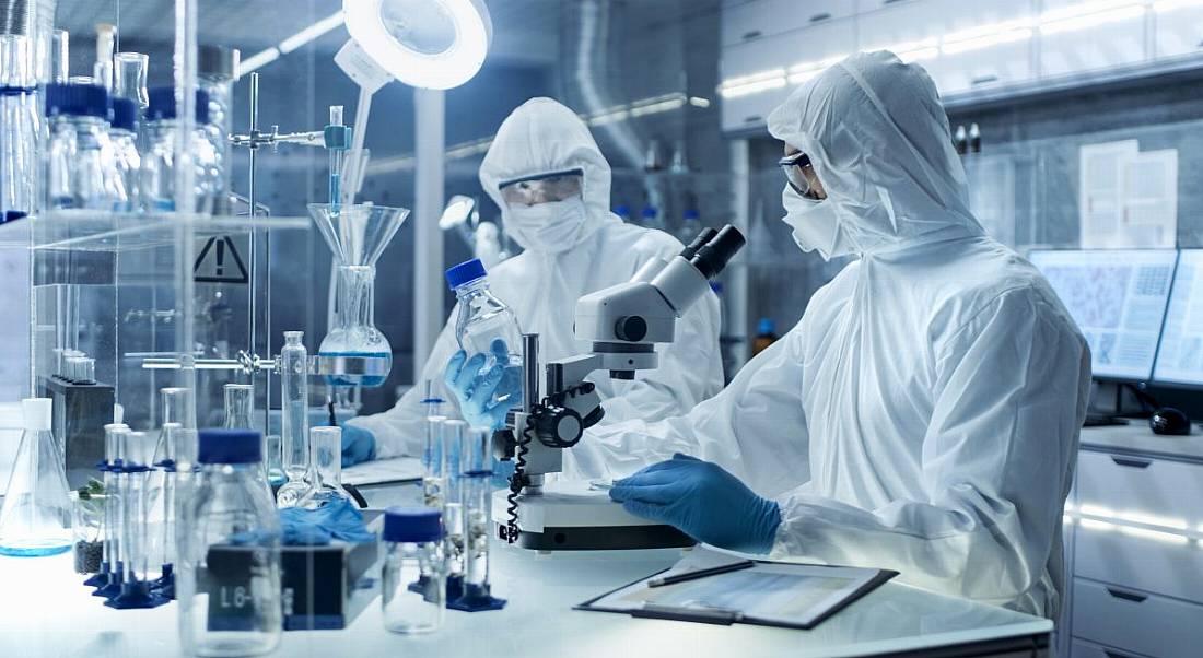 Two scientists in full protective gear work in a lab with a microscope and other biopharma paraphernalia in front of them.