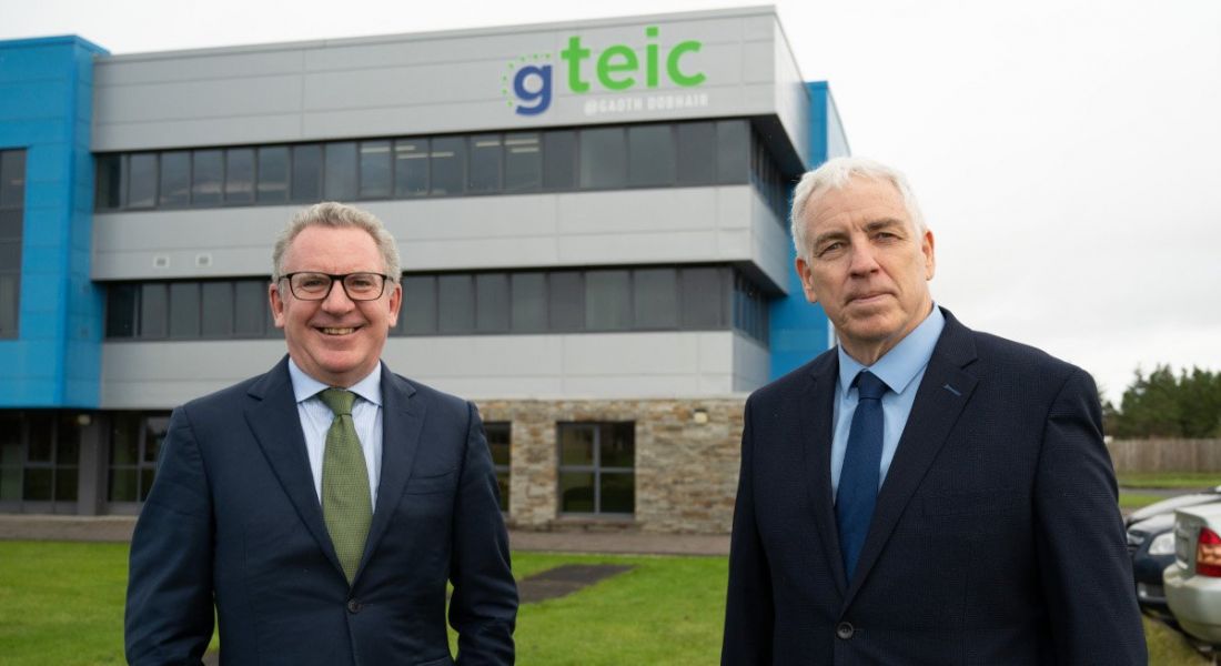 Two men in suits stand outside a large building that says Gteic on the side of it.