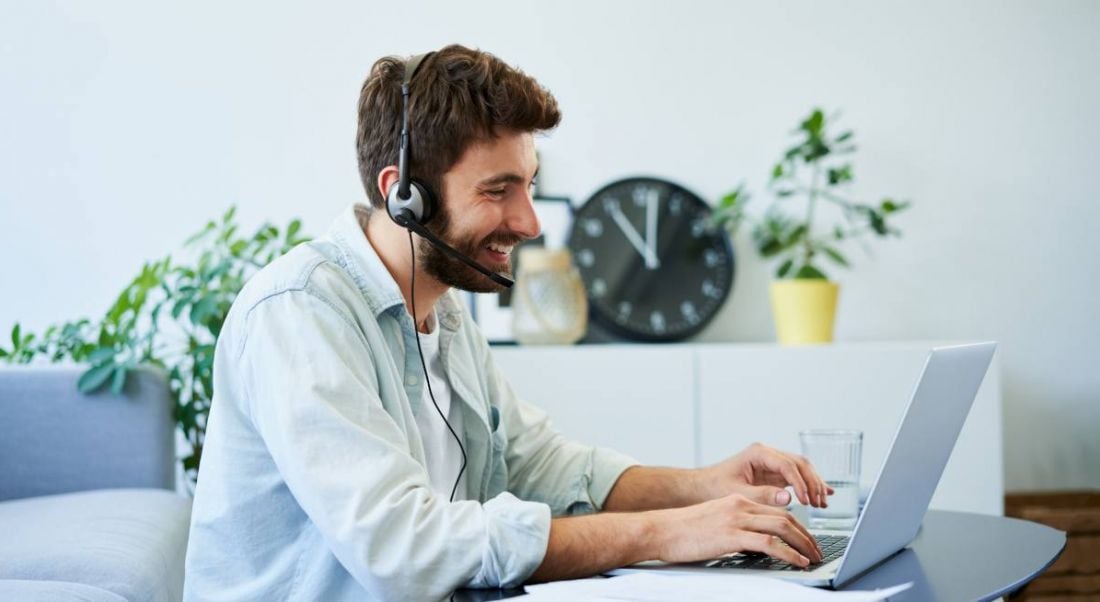 A young man wearing a casual shirt in a bright room is wearing a headset and working at a laptop, symbolising hybrid working.