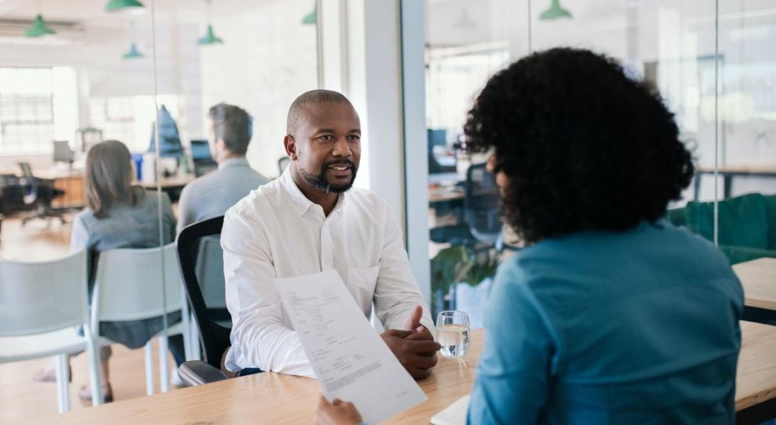 A woman holding a CV sitting opposite a man in a white shirt in an interview setting in a bright office.