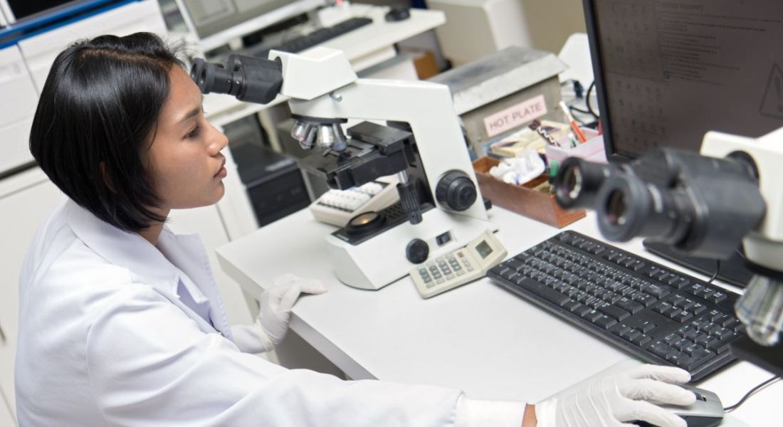 Woman in a white coat working in a lab on a computer. There is a microscope beside her.