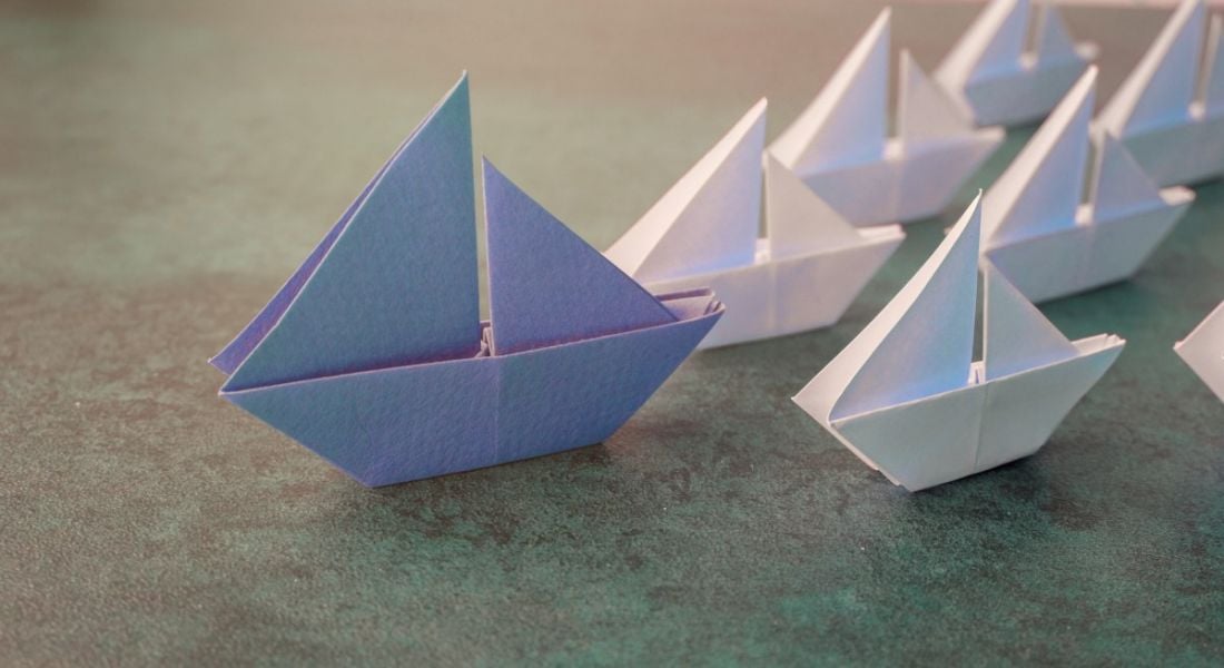 A large origami blue paper boat leads several smaller white paper boats on a greenish surface, symbolising tech leaders.