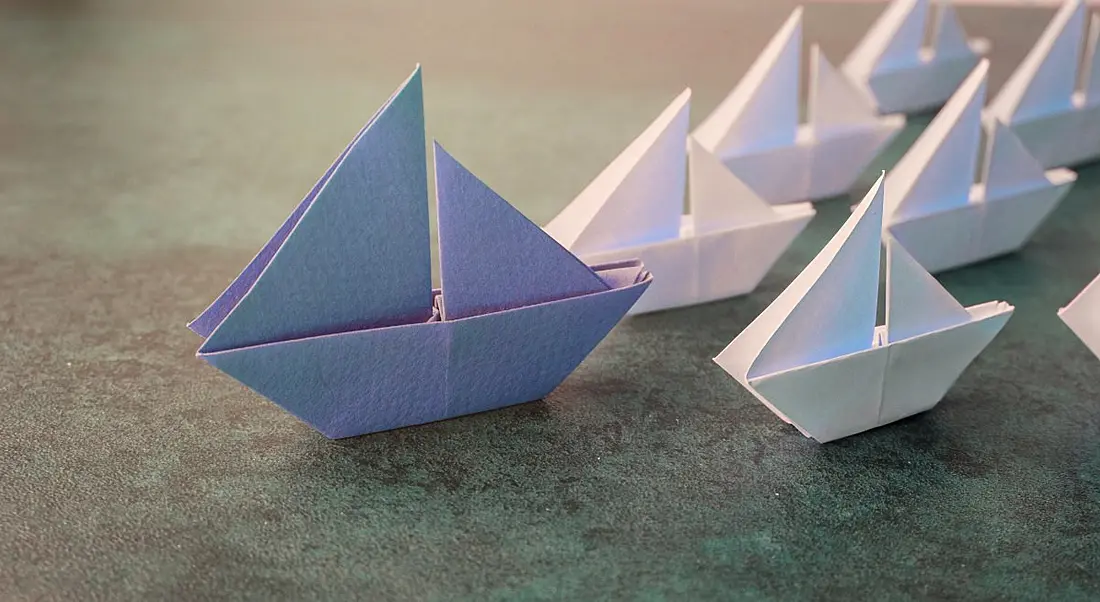 A large origami blue paper boat leads several smaller white paper boats on a greenish surface, symbolising tech leaders.