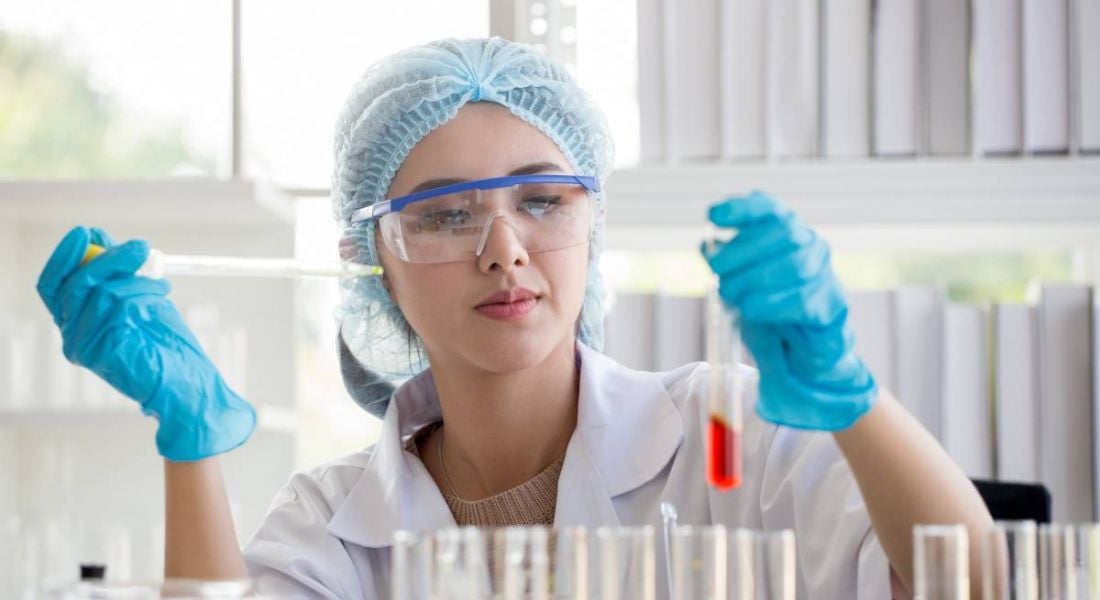 A woman in a white lab coat and protective eyewear and gloves holds a pipette and a test tube to depict working in pharma.