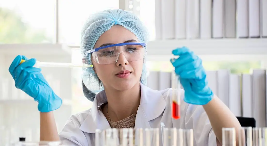 A woman in a white lab coat and protective eyewear and gloves holds a pipette and a test tube to depict working in pharma.