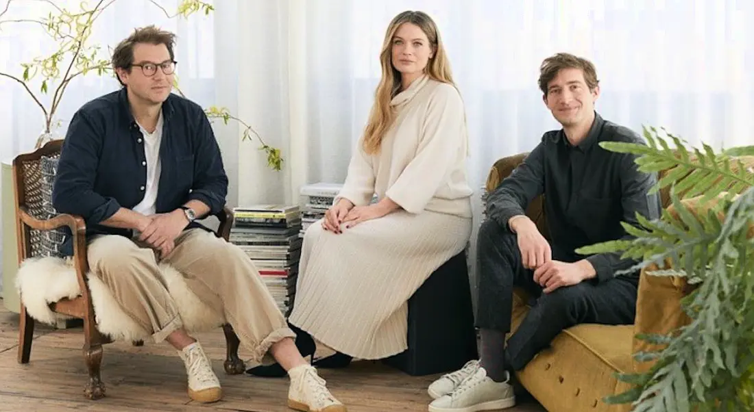 The three founders of &Open sitting in a casual office setting on sofas surrounded by plants and modern, neutral furnishings.