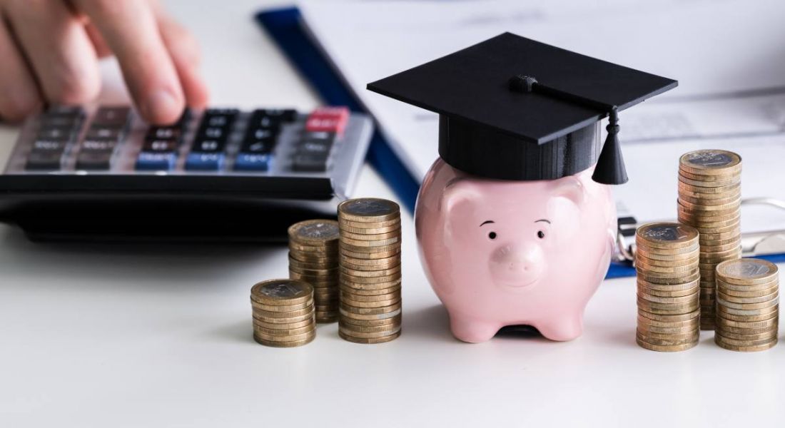 Piggy bank wearing a graduation cap beside stacks of coins and a calculator and clipboard.