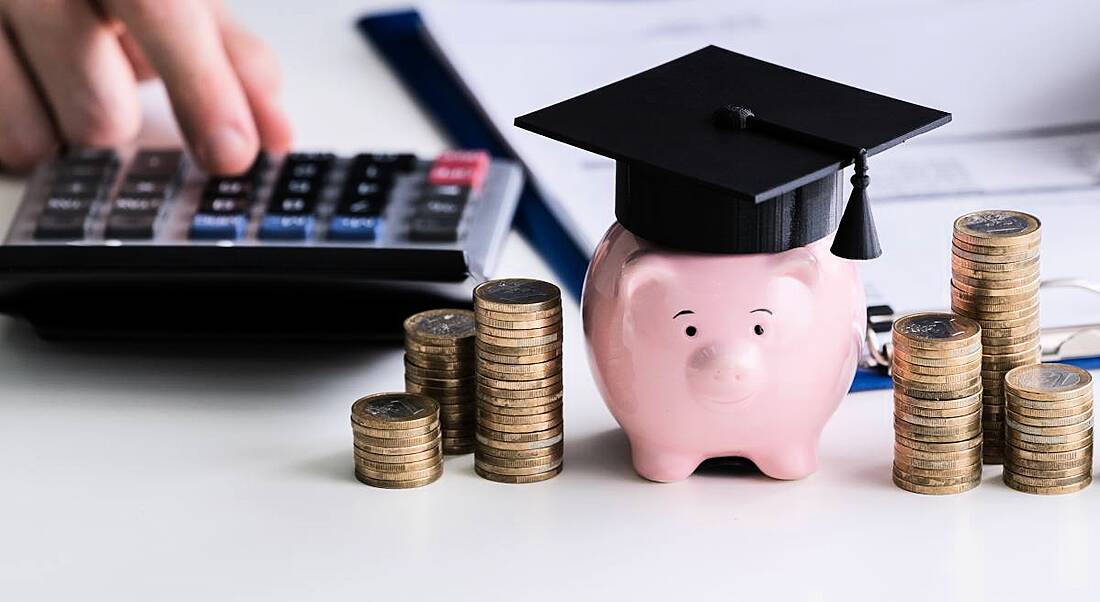 Piggy bank wearing a graduation cap beside stacks of coins and a calculator and clipboard.