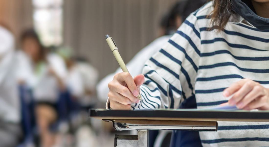 Student holding a pen while taking an exam on a wooden desk, with more people taking the exam in the background.