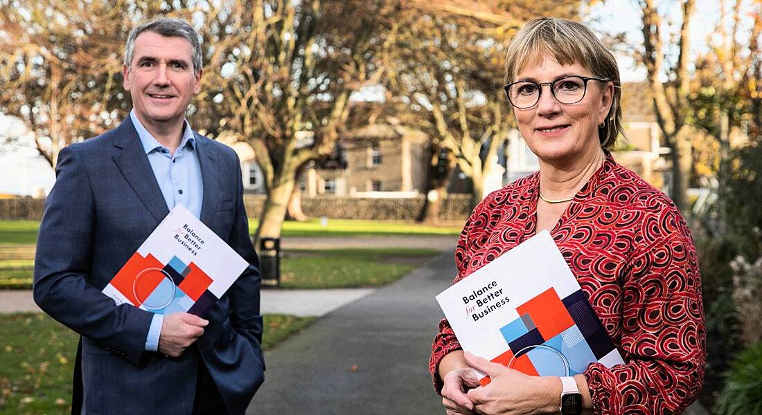 Aongus Hegarty and Julie Sinnamon holding the Balance For Better Business report standing outside on a tree-lined avenue.