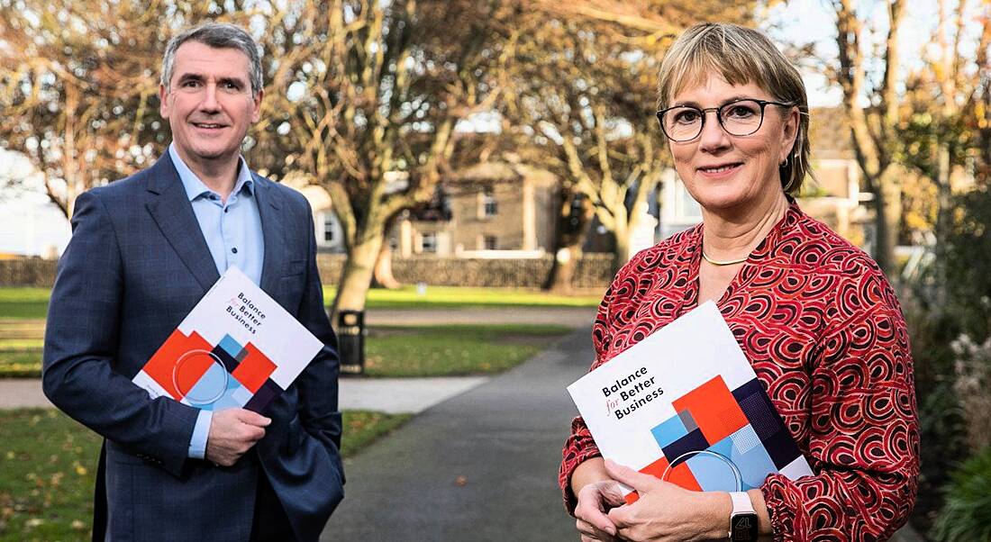 Aongus Hegarty and Julie Sinnamon holding the Balance For Better Business report standing outside on a tree-lined avenue.