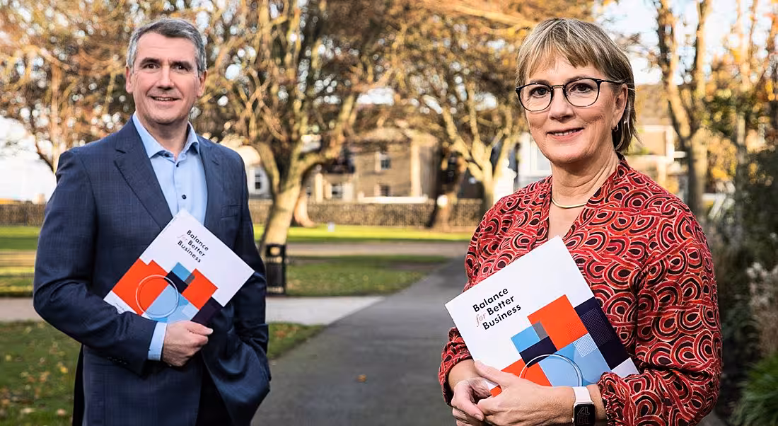 Aongus Hegarty and Julie Sinnamon holding the Balance For Better Business report standing outside on a tree-lined avenue.